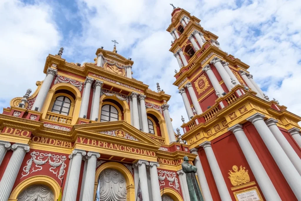De opvallende rood-gouden gevel en de hoge klokkentoren van de San Francisco-basiliek in Salta, gezien vanuit een laag perspectief tegen een bewolkte lucht.