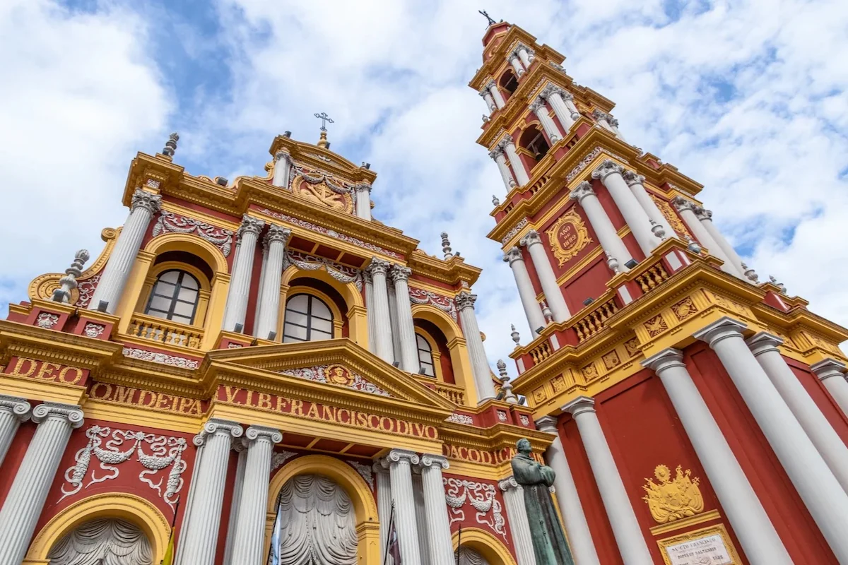 De opvallende rood-gouden gevel en de hoge klokkentoren van de San Francisco-basiliek in Salta, gezien vanuit een laag perspectief tegen een bewolkte lucht.