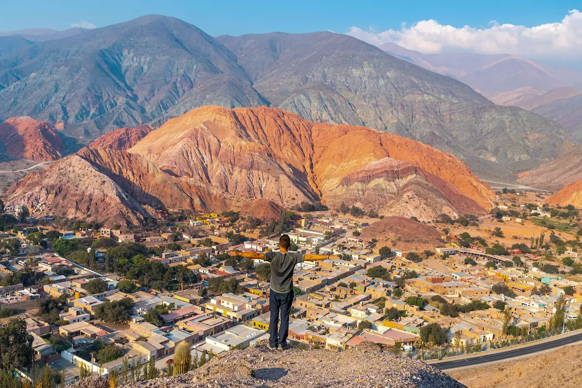 Een man staat met gespreide armen en kijkt uit over het Andesdorp Purmamarca met de indrukwekkende Berg van de Zeven Kleuren (Cerro de los Siete Colores) op de achtergrond.