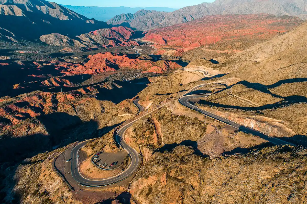 Een spectaculair luchtpanorama van de slingerende haarspeldbochten van de Cuesta de Miranda op de Ruta 40, omgeven door dieprode en bruine berglandschappen.