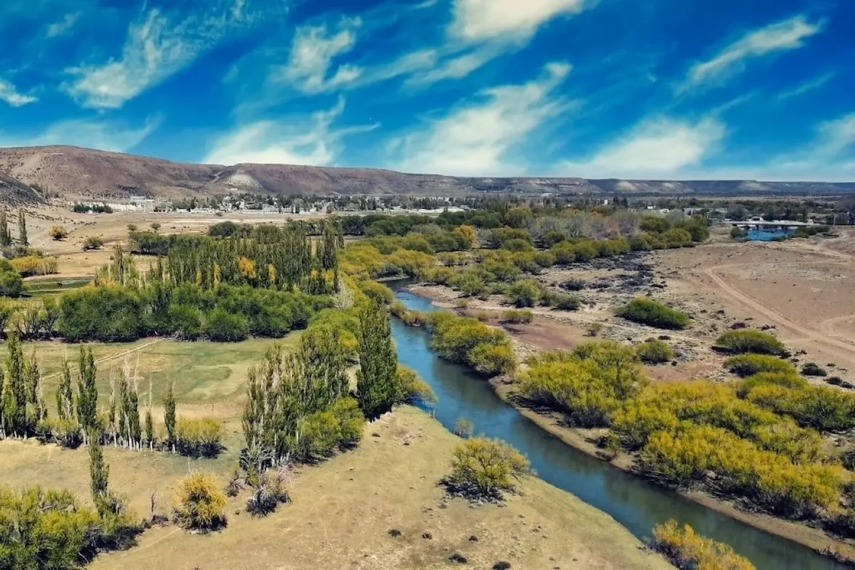 Een weids uitzicht over de goudgele Patagonische steppe met een kronkelende rivier en verre bergen onder een dynamische wolkenlucht bij Río Mayo.