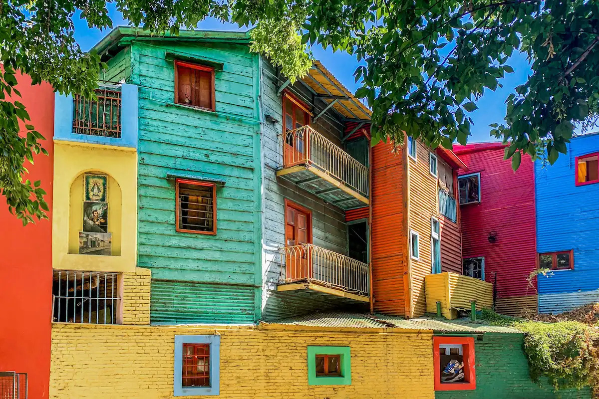 De iconische, bontgekleurde huisjes van golfplaat en hout in de straat El Caminito in de wijk La Boca, Buenos Aires, gezien door een frame van groene bladeren.