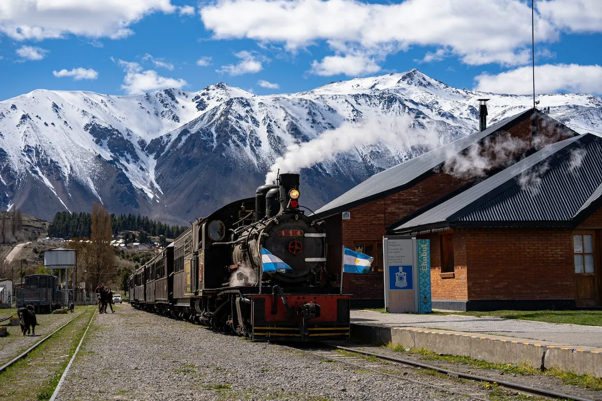 De historische stoomtrein La Trochita bij het station in Esquel met op de achtergrond de besneeuwde toppen van de Andes onder een blauwe lucht.