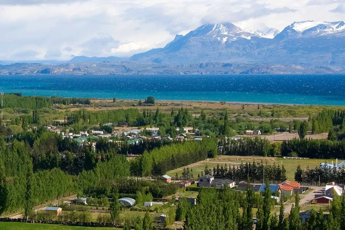 Zicht op het uitgestrekte blauwe Lago Buenos Aires bij Los Antiguos met groene populieren op de voorgrond en witte Andes toppen in de verte.