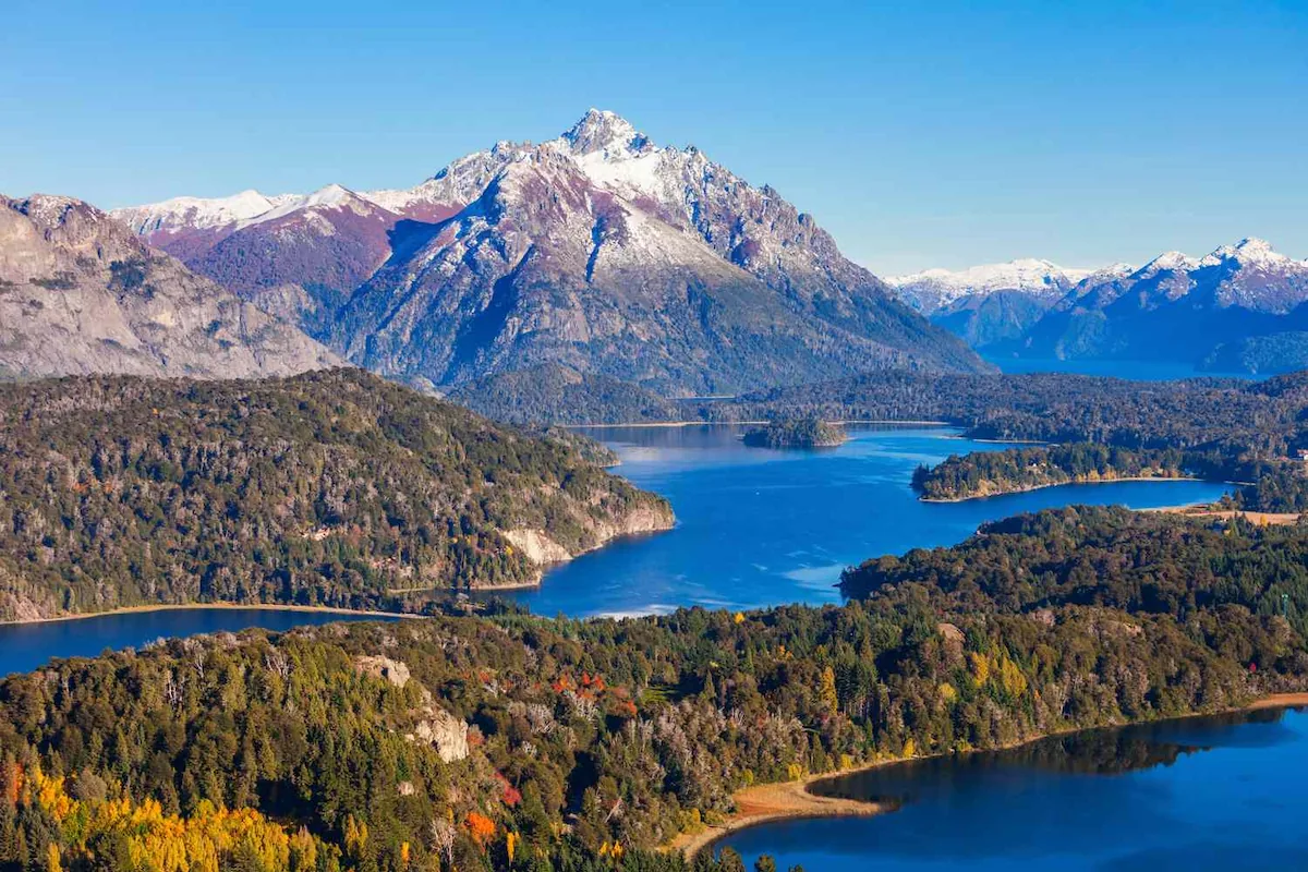 Een weids uitzicht over de diepblauwe meren en beboste eilanden van Bariloche met besneeuwde witte Andes toppen op de achtergrond.