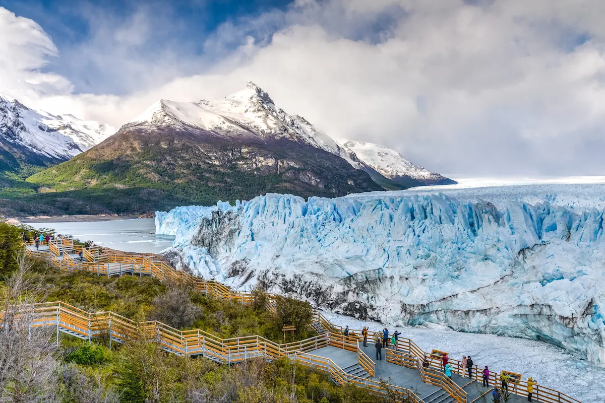 Panoramisch uitzicht op de Perito Moreno gletsjer vanaf de houten loopbruggen in het Nationaal Park Los Glaciares, met besneeuwde bergtoppen en toeristen op de uitkijkpunten.