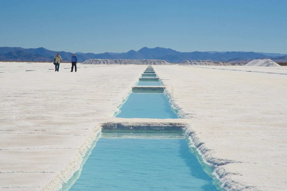 Helderblauwe waterkanalen snijden door de uitgestrekte witte zoutvlakte van de Salinas Grandes in Noord-Argentinië, met bergen aan de horizon.