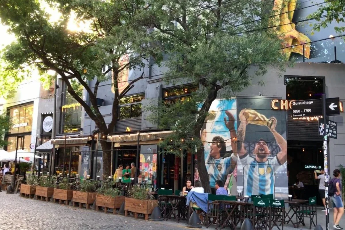 Mensen op een terras in de wijk Palermo Soho in Buenos Aires, naast een grote muurschildering van voetbaliconen Maradona en Messi.