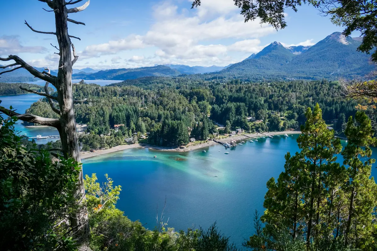 Een panoramisch uitzicht op de diepblauwe baai Bahía Mansa in Villa La Angostura, omringd door dichte groene bossen en de toppen van de Andes.