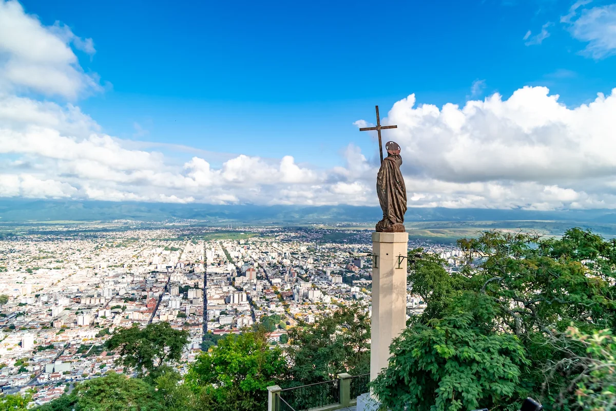 Panoramisch uitzicht over de stad Salta vanaf de top van de Cerro San Bernardo, met een religieus standbeeld op de voorgrond en de Andes in de verte.