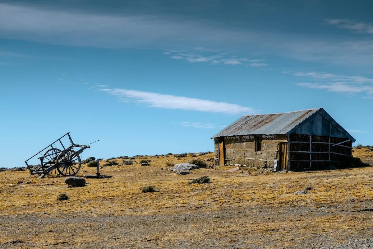 Een rustiek stenen schuurtje met een golfplaten dak en een antieke houten kar op de dorre Patagonische steppe onder een helderblauwe lucht.