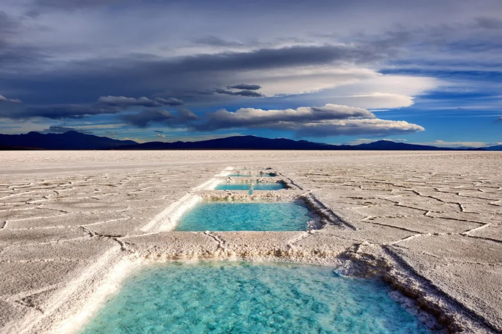 Turquoise waterbassins in de witte zoutvlakte van Salinas Grandes onder een indrukwekkende bewolkte lucht met bergen aan de horizon.