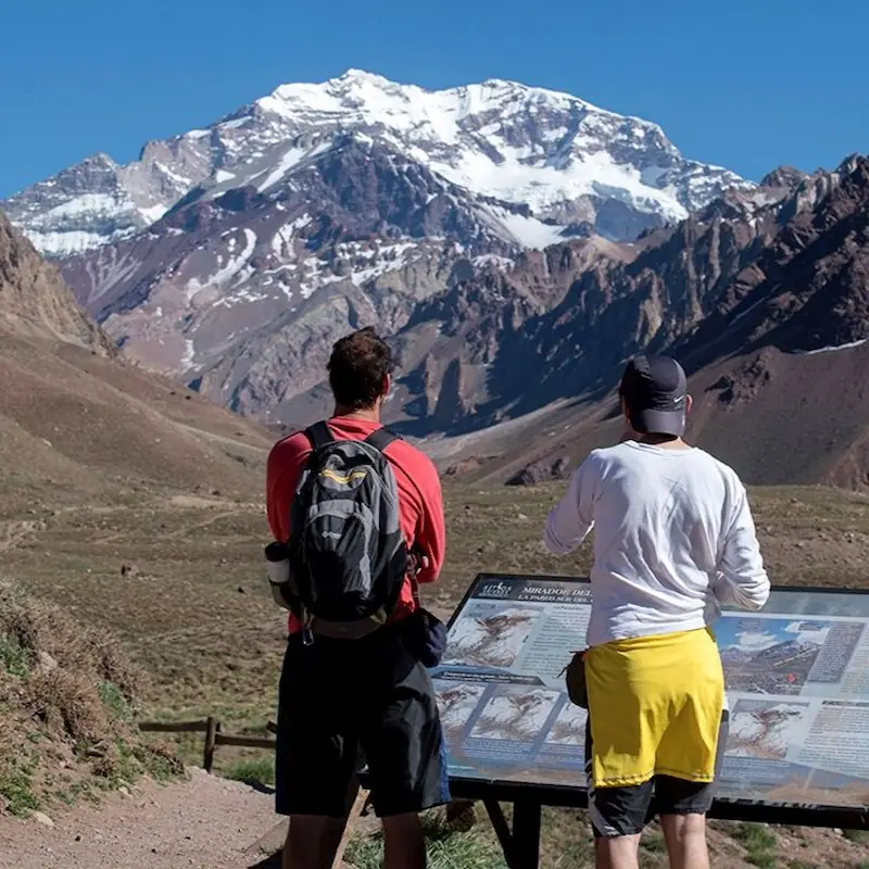 Twee reizigers bij een informatiebord kijken uit op de besneeuwde zuidwand van de Aconcagua in het Andesgebergte.