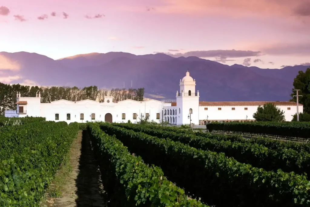De wijngaarden en het witte koloniale gebouw van Bodega El Esteco in Cafayate, Salta, tegen een achtergrond van bergen bij zonsondergang.
